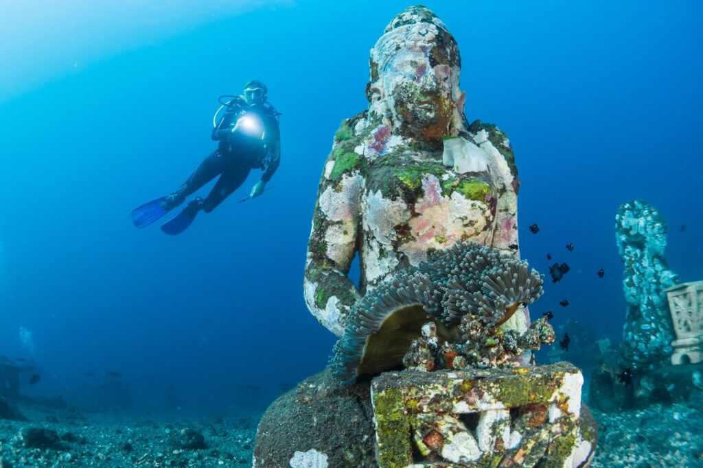 Buceador con linterna tras una estatua sumergida cubierta de coral, con una anémona en el regazo