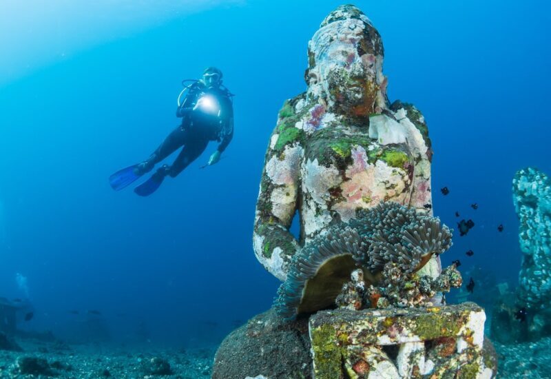 Buceador con linterna tras una estatua sumergida cubierta de coral, con una anémona en el regazo